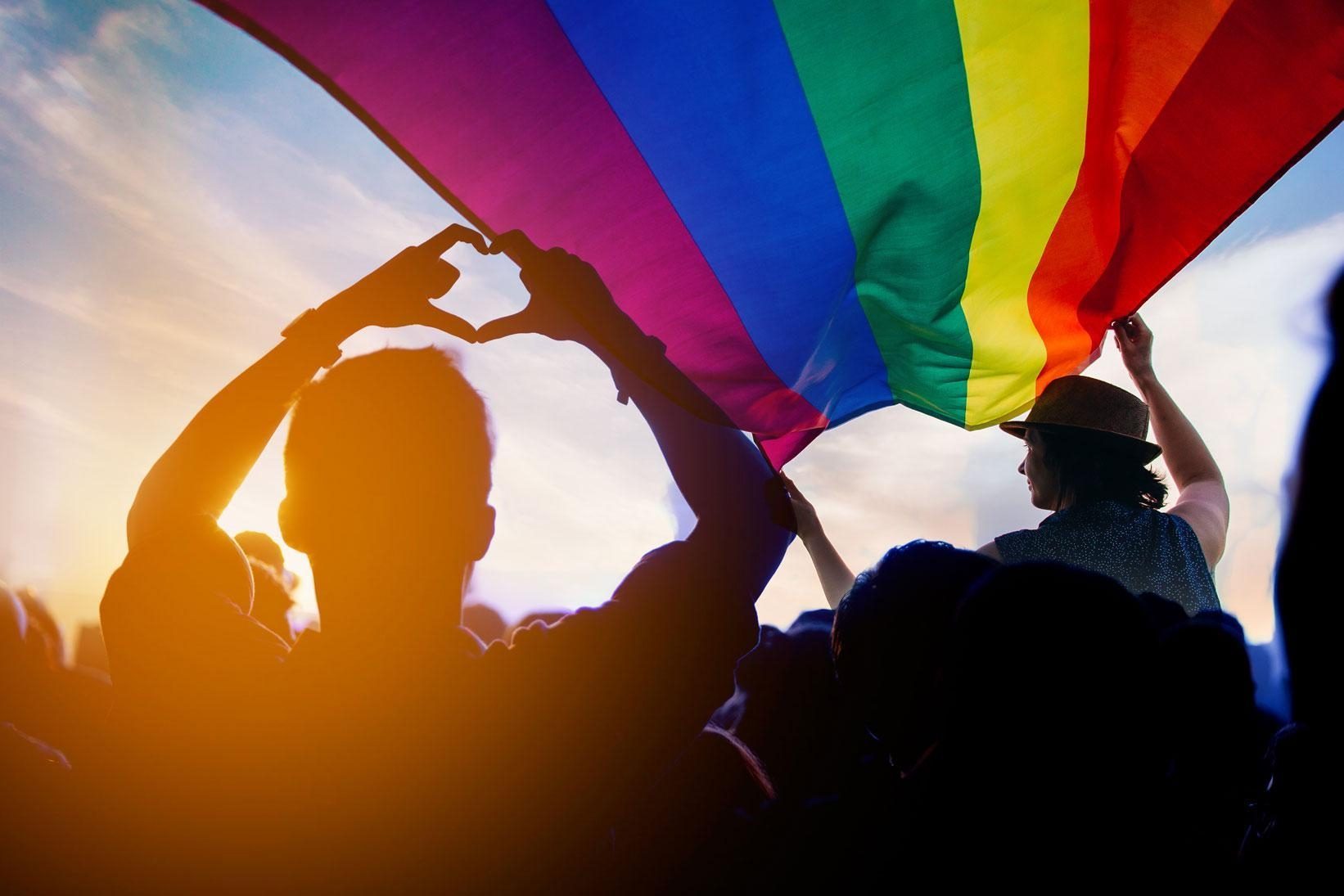 Une personne forme un coeur avec ses doigts devant un drapeau arc-en-ciel